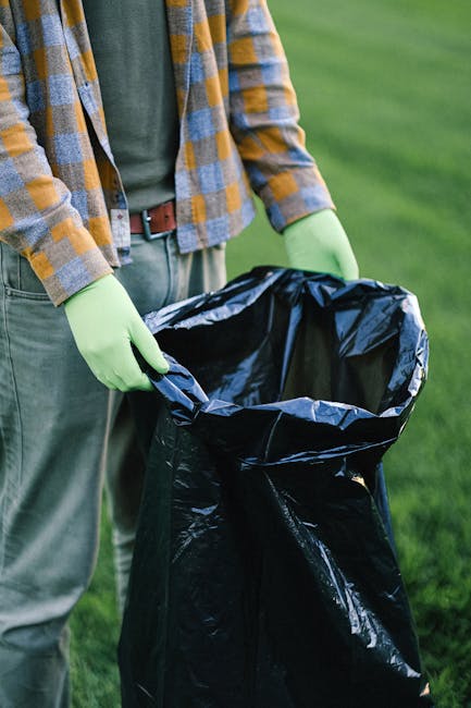 A person standing outdoors on a grassy area, wearing a yellow and gray checkered jacket, green gloves, and casual clothing. They are holding open a large black rubbish bag made of plastic, which appea